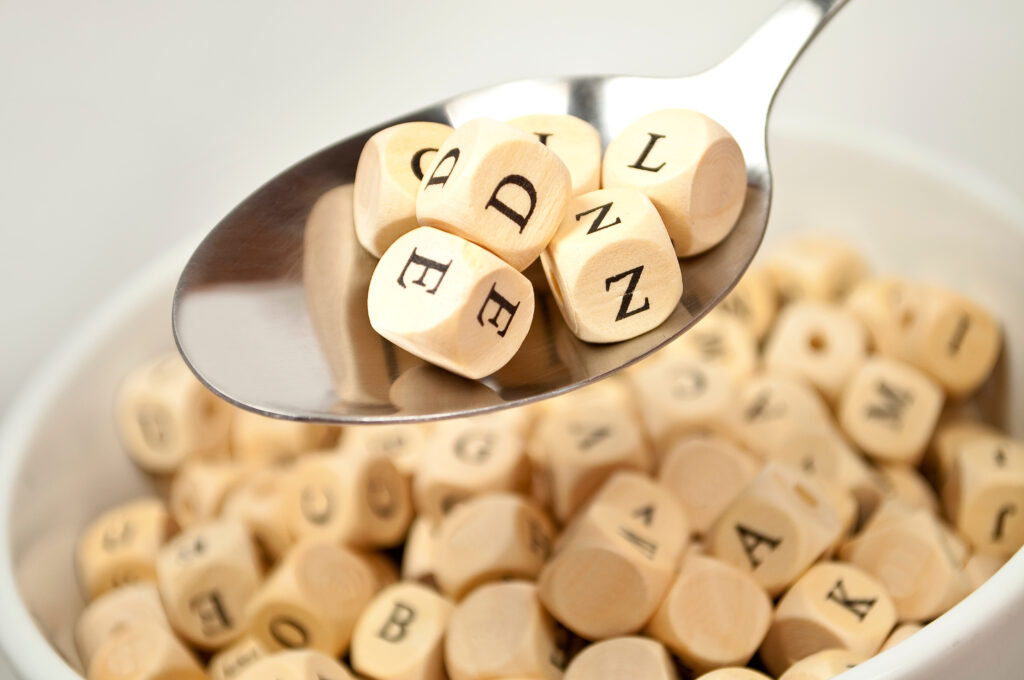 Buchstabenwürfel in einer Tasse, darüber ein Löffel mit ein paar Würfeln